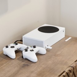 White Xbox Series S with two white controllers resting on a wooden table, plugged in against a neutral wall.