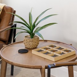 Round wooden coffee table with a green plant in a woven basket and a wooden grid-style game beside a brown leather chair.