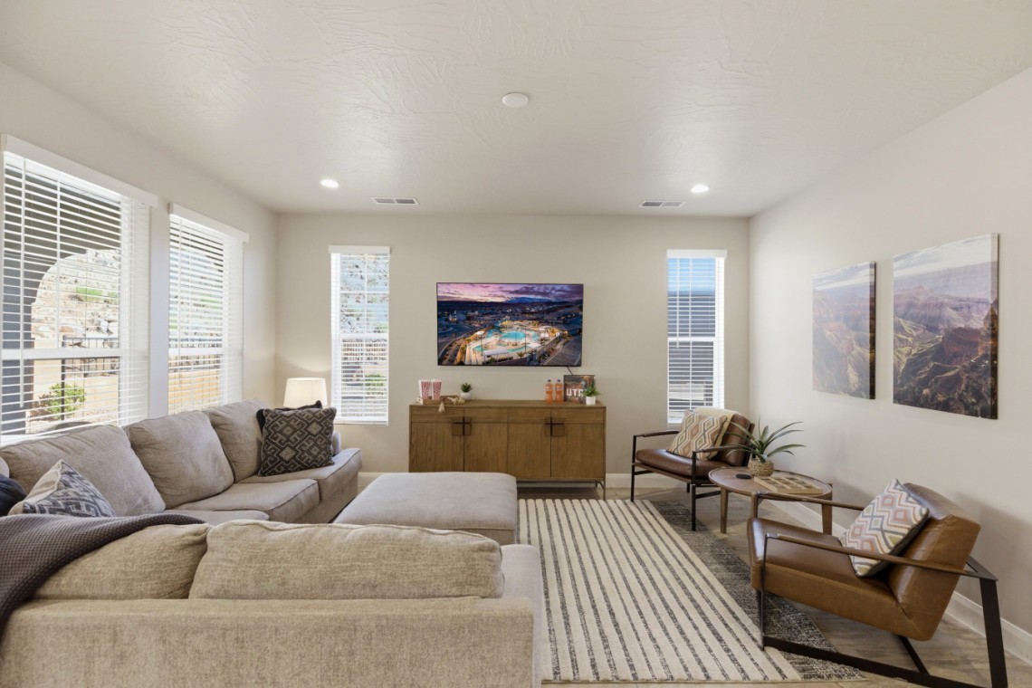 Bright living room with a beige sectional, striped rug, two brown armchairs, and a wall-mounted TV above a wooden console.