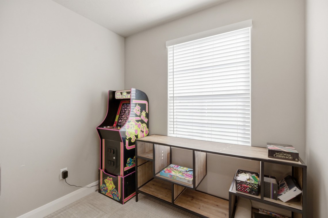 A small game corner with a vintage arcade cabinet beside a long wooden desk and shelving under a window with white blinds.