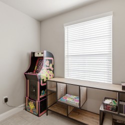 A small game corner with a vintage arcade cabinet beside a long wooden desk and shelving under a window with white blinds.