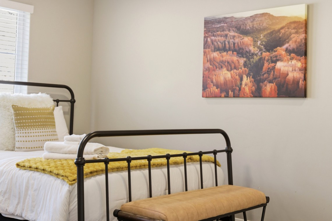 Metal bed frame with white linens, a yellow knit throw, and folded towels on a bench at the foot of the bed in a bright bedroom, with a canyon landscape painting on the wall.