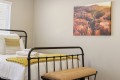 Metal bed frame with white linens, a yellow knit throw, and folded towels on a bench at the foot of the bed in a bright bedroom, with a canyon landscape painting on the wall.