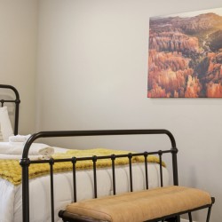 Metal bed frame with white linens, a yellow knit throw, and folded towels on a bench at the foot of the bed in a bright bedroom, with a canyon landscape painting on the wall.