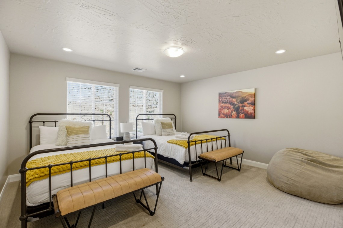 Two twin metal-frame beds with white linens and bright yellow knit throws, matching benches at the foot, in a neutral-lit bedroom with two windows and a landscape print on the wall.