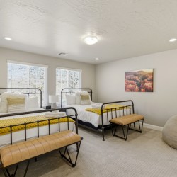 Two twin metal-frame beds with white linens and bright yellow knit throws, matching benches at the foot, in a neutral-lit bedroom with two windows and a landscape print on the wall.