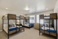Eight-bed dorm room with metal-framed bunk beds, wood drawer panels, and blue bedding against beige walls and carpet.