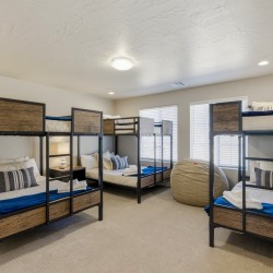 Eight-bed dorm room with metal-framed bunk beds, wood drawer panels, and blue bedding against beige walls and carpet.