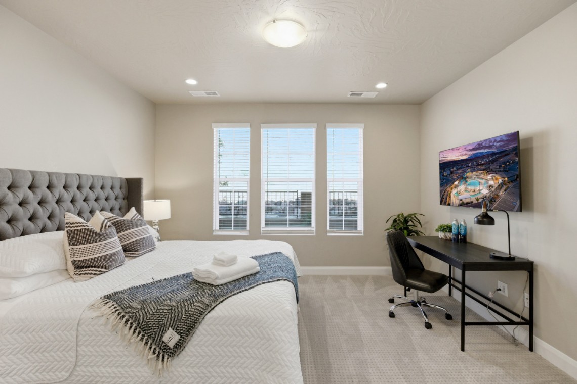 Bedroom with a large tufted headboard, white quilted bed, and decorative pillows; a gray throw blanket sits at the foot of the bed, towels on top. A desk with a monitor and a chair is against the right wall, with a lamp and small plants nearby. Three tall windows let in natural light.