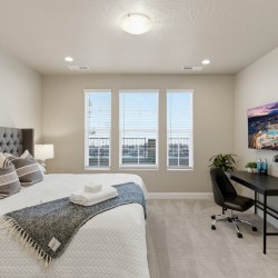 Bedroom with a large tufted headboard, white quilted bed, and decorative pillows; a gray throw blanket sits at the foot of the bed, towels on top. A desk with a monitor and a chair is against the right wall, with a lamp and small plants nearby. Three tall windows let in natural light.