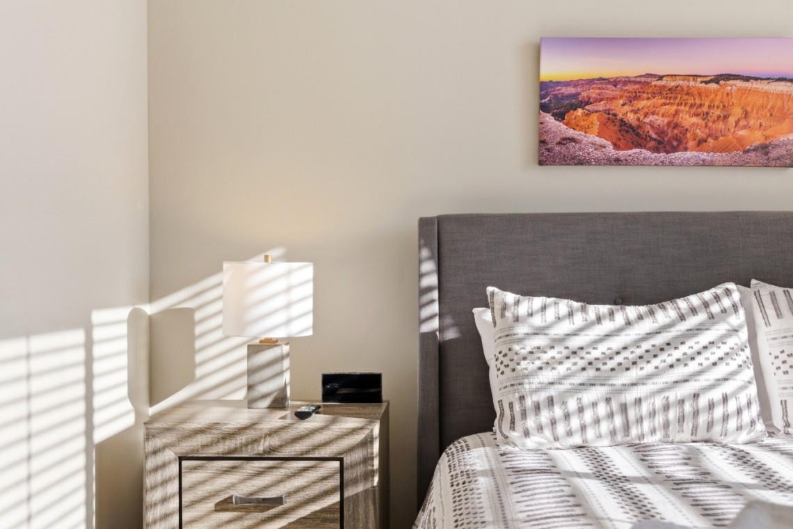 Modern bedroom with gray upholstered headboard, white patterned duvet, nightstand, lamp, and a landscape photo above the bed in sunset tones.