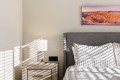 Modern bedroom with gray upholstered headboard, white patterned duvet, nightstand, lamp, and a landscape photo above the bed in sunset tones.