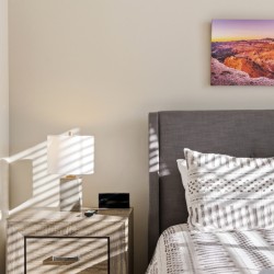 Modern bedroom with gray upholstered headboard, white patterned duvet, nightstand, lamp, and a landscape photo above the bed in sunset tones.