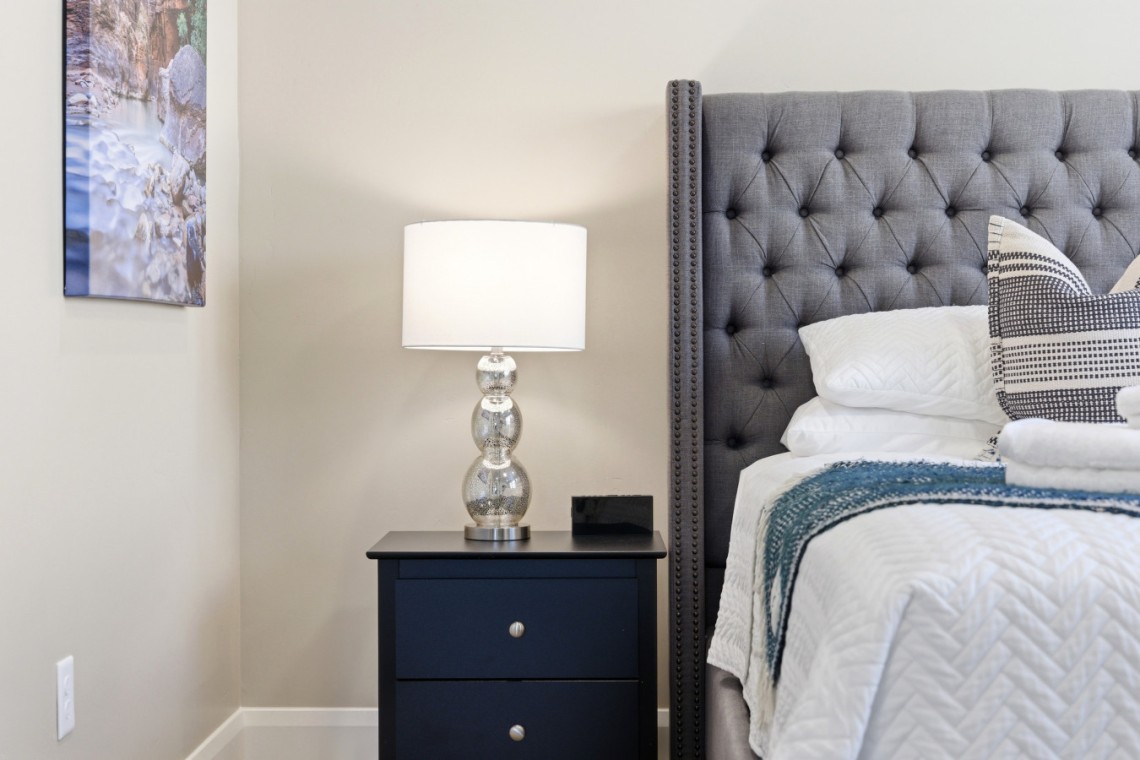 Nightstand with a glass-lamp beside a tufted gray headboard bed and white bedding in a cozy bedroom corner.