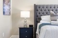 Nightstand with a glass-lamp beside a tufted gray headboard bed and white bedding in a cozy bedroom corner.