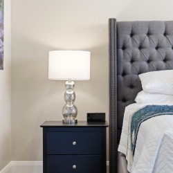 Nightstand with a glass-lamp beside a tufted gray headboard bed and white bedding in a cozy bedroom corner.
