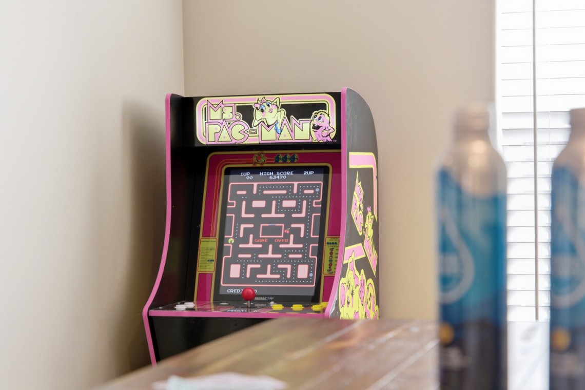 Pink Ms. Pac-Man arcade cabinet against a beige wall, with the game screen on and a wooden table in the foreground; two blue bottles are blurred to the right.