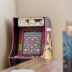 Pink Ms. Pac-Man arcade cabinet against a beige wall, with the game screen on and a wooden table in the foreground; two blue bottles are blurred to the right.