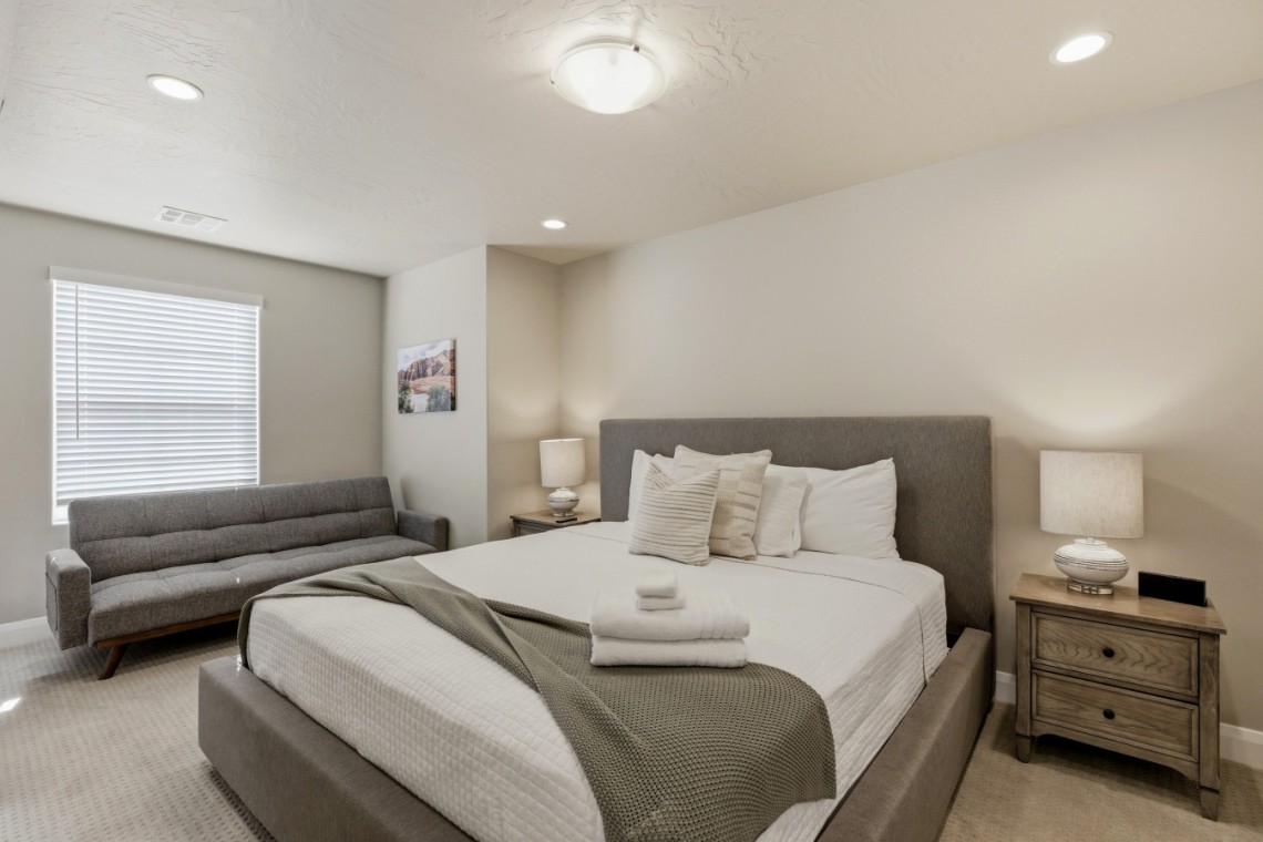 Hotel-style bedroom with a gray upholstered king bed, matching headboard, two nightstands, and a gray sofa against the wall at left.