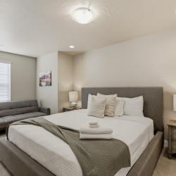 Hotel-style bedroom with a gray upholstered king bed, matching headboard, two nightstands, and a gray sofa against the wall at left.