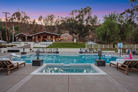Resort-style pool area with lounge chairs, umbrellas, and a clubhouse in the background at sunset.