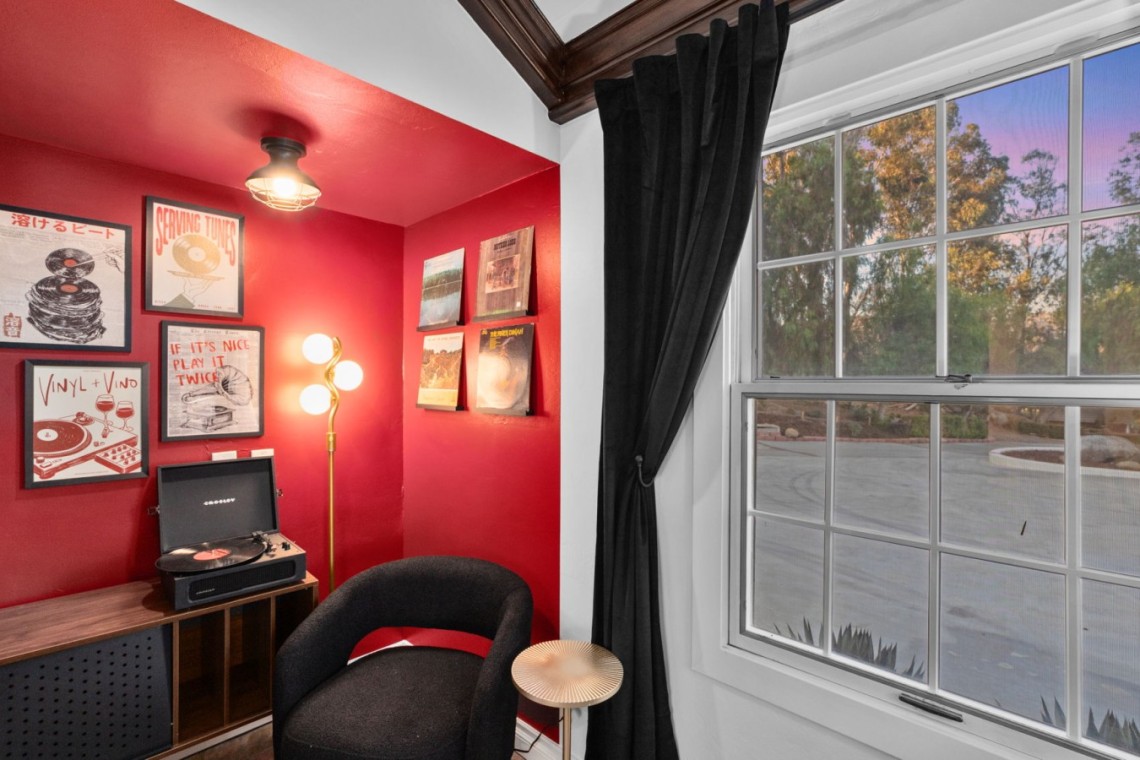 Cozy red-walled listening nook with a turntable on a wooden cabinet, framed vinyl posters, and a black curved chair by a large window with dark curtains.