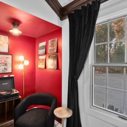 Cozy red-walled listening nook with a turntable on a wooden cabinet, framed vinyl posters, and a black curved chair by a large window with dark curtains.