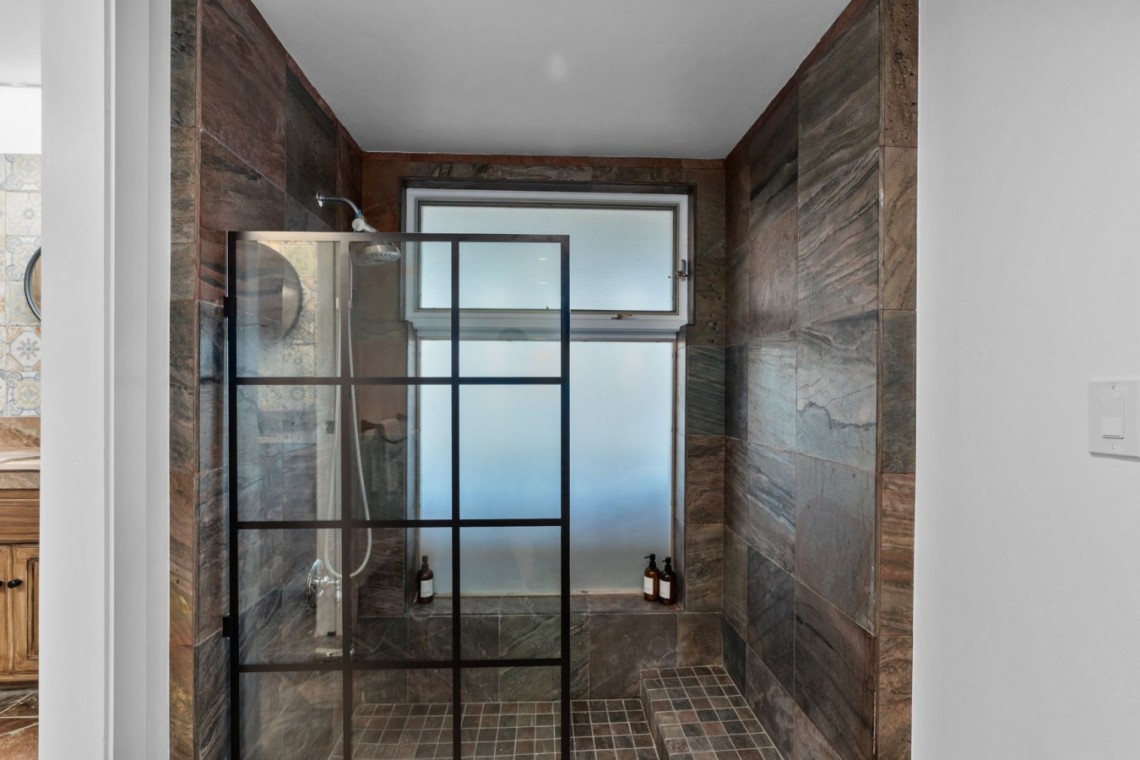 Walk-in shower with a black-framed glass door, dark stone walls, and a frosted window; a few bottles sit on the shower ledge/corner shelf.