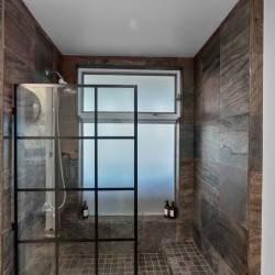 Walk-in shower with a black-framed glass door, dark stone walls, and a frosted window; a few bottles sit on the shower ledge/corner shelf.