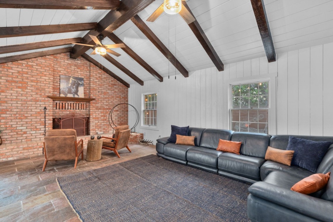 Living room with an exposed brick fireplace, dark wooden ceiling beams, and a large dark leather sectional sofa with orange and navy cushions.