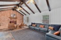 Living room with an exposed brick fireplace, dark wooden ceiling beams, and a large dark leather sectional sofa with orange and navy cushions.