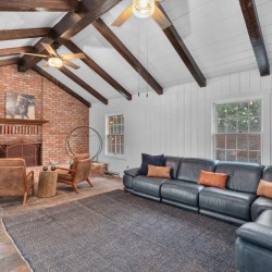 Living room with an exposed brick fireplace, dark wooden ceiling beams, and a large dark leather sectional sofa with orange and navy cushions.