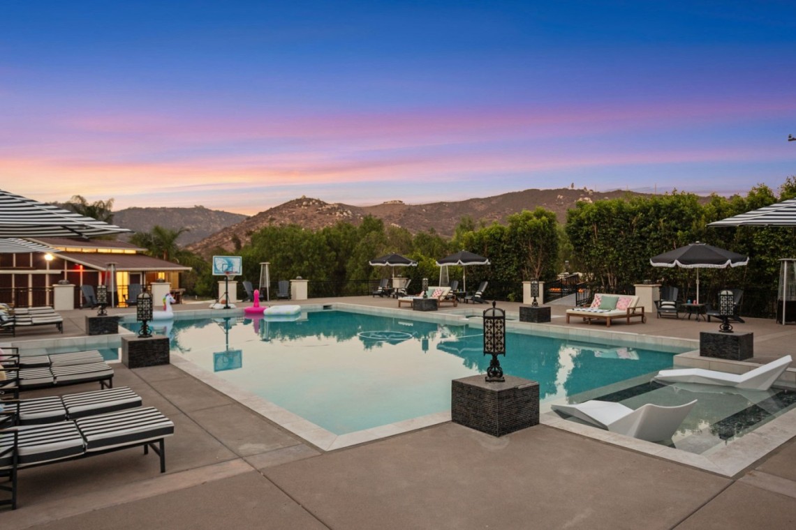 Luxurious outdoor pool area at dusk with striped lounge chairs, umbrellas, and mountain backdrop.