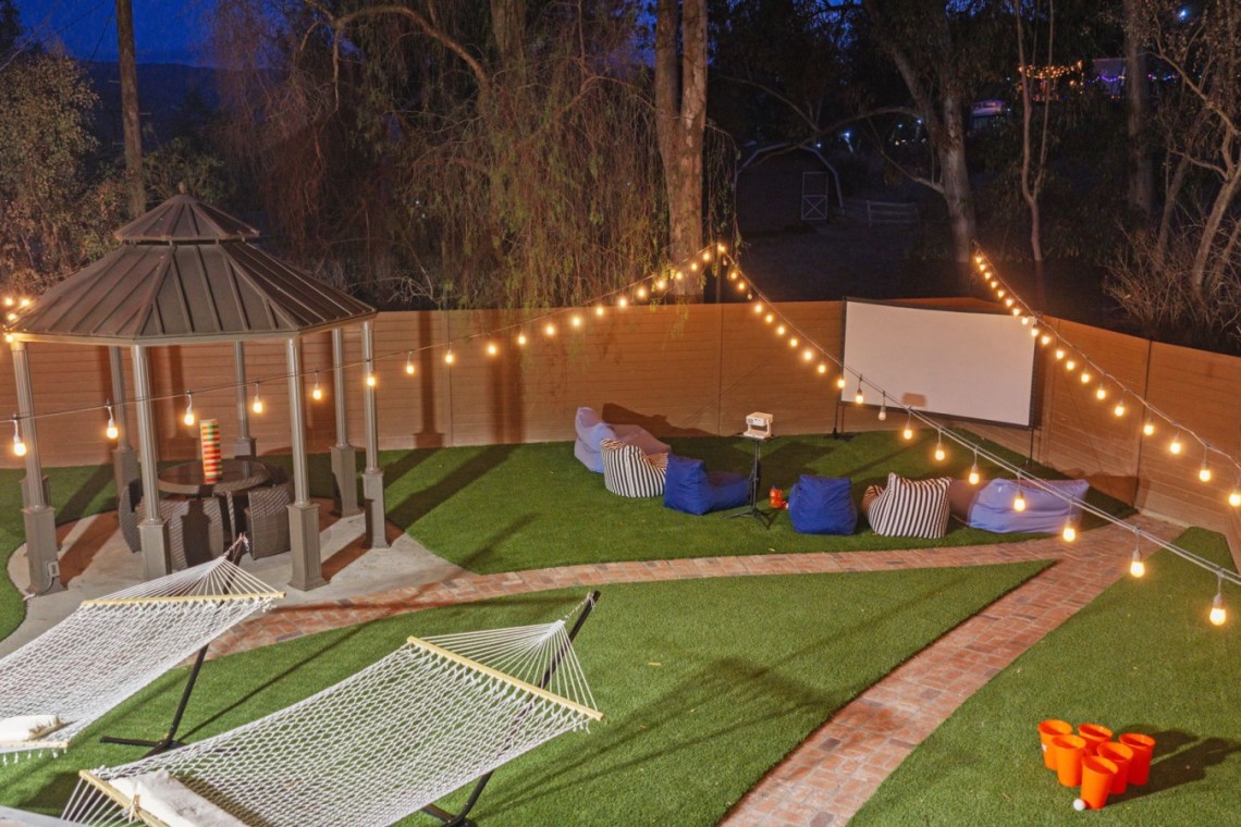Nighttime backyard party setup with string lights, a central gazebo, hammocks, and a projector screen.