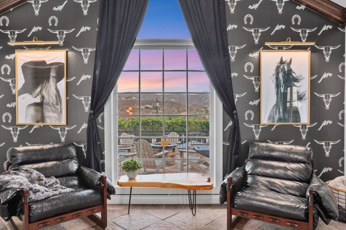 Cozy seating area with two black leather chairs facing a large window showing a sunset over a patio, framed horse and figure portraits, and horse-themed wallpaper.