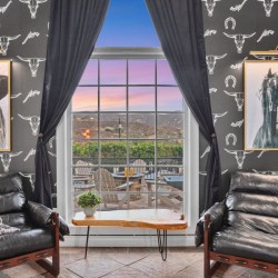 Cozy seating area with two black leather chairs facing a large window showing a sunset over a patio, framed horse and figure portraits, and horse-themed wallpaper.