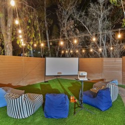 Outdoor backyard movie setup at night with a white projection screen, projector on a stand, string lights overhead, and blue and striped bean bag chairs on artificial turf.