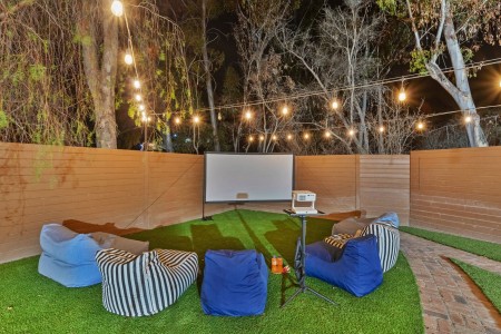Outdoor backyard movie setup at night with a white projection screen, projector on a stand, string lights overhead, and blue and striped bean bag chairs on artificial turf.