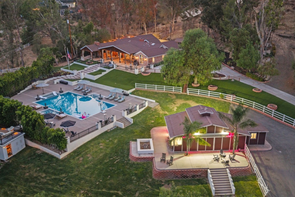 Aerial view of a luxury estate with a rectangular pool, lounge chairs, umbrellas, and a clubhouse surrounded by lawns and trees, at dusk light.”,