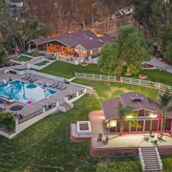 Aerial view of a luxury estate with a rectangular pool, lounge chairs, umbrellas, and a clubhouse surrounded by lawns and trees, at dusk light.”,