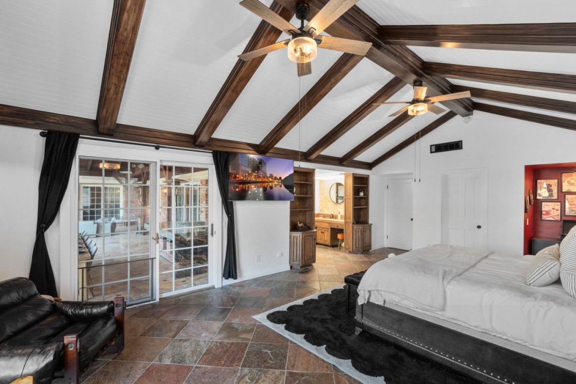 Bedroom with vaulted ceiling and dark wood beams, a king bed on the right, and glass doors opening to a patio. Pristine white walls and a red accent wall with art on the far side, plus ceiling fans overhead.