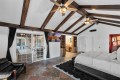 Bedroom with vaulted ceiling and dark wood beams, a king bed on the right, and glass doors opening to a patio. Pristine white walls and a red accent wall with art on the far side, plus ceiling fans overhead.
