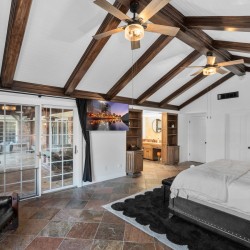 Bedroom with vaulted ceiling and dark wood beams, a king bed on the right, and glass doors opening to a patio. Pristine white walls and a red accent wall with art on the far side, plus ceiling fans overhead.