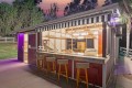 Outdoor night bar with a striped awning, marble counter, and three wooden stools; interior reveals a pool table and seating area.