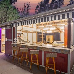 Outdoor night bar with a striped awning, marble counter, and three wooden stools; interior reveals a pool table and seating area.