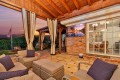Cozy covered patio with beige seating and purple cushions, brick wall, and a TV mounted outside at sunset over green landscape.