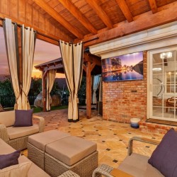 Cozy covered patio with beige seating and purple cushions, brick wall, and a TV mounted outside at sunset over green landscape.