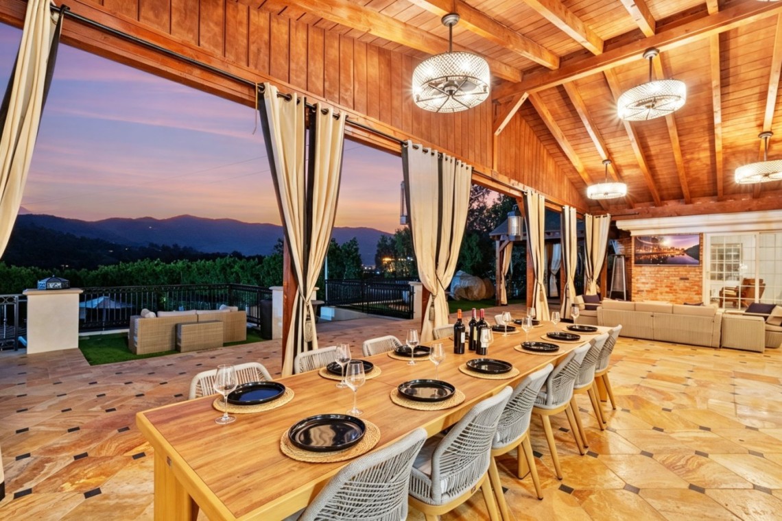 Long wooden dining table set for a meal in a covered patio, with wine bottles, place settings, and cream curtains, overlooking a sunset mountain view.