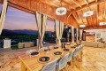 Long wooden dining table set for a meal in a covered patio, with wine bottles, place settings, and cream curtains, overlooking a sunset mountain view.