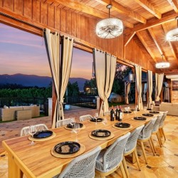 Long wooden dining table set for a meal in a covered patio, with wine bottles, place settings, and cream curtains, overlooking a sunset mountain view.
